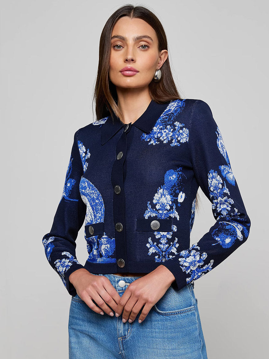 A woman in the L'Agence Osiris Jacquard Cardigan in Black/Blue Large Porcelain and light blue jeans stands against a plain gray background.