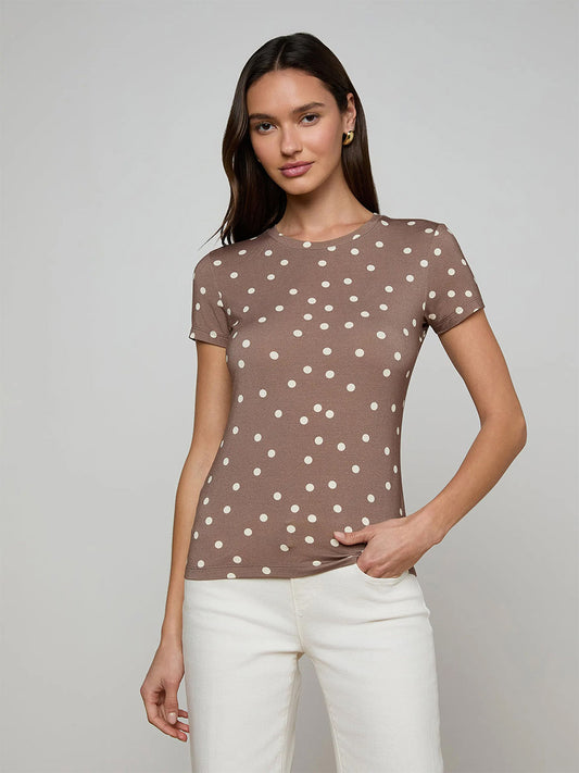 A woman stands against a plain background, wearing the L'Agence Ressi Fitted Tee in Cardamom/Ecru Polka Dot—bestselling, slim cut with brown short sleeves and white polka dots—paired with white pants.