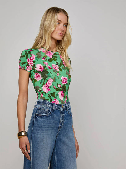 A woman with long blonde hair wears the L'Agence Ressi Fitted Tee in Green Multi Santa Fe Rose and high-waisted blue jeans, standing against a plain light background.