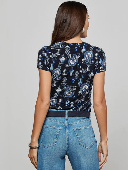 A woman with long brown hair, wearing the L'Agence Ressi Fitted Tee in Black/Blue Porcelain Print and high-waisted blue jeans, stands facing away from the camera.