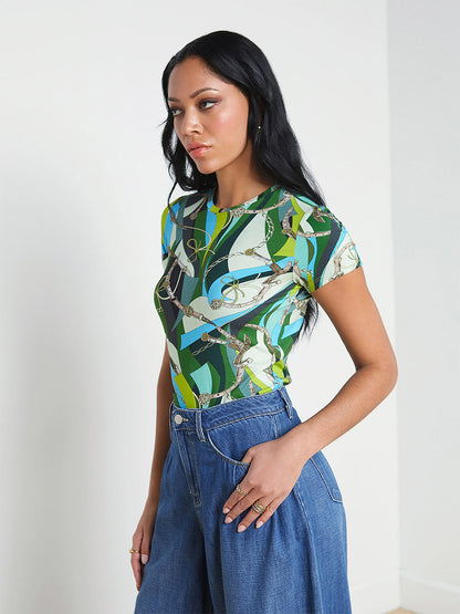 A woman with a dark complexion stands in a white room, wearing a L'Agence Ressi Fitted Tee in Small Sea Green Belt Swirl top and blue jeans, with her hand in her pocket.