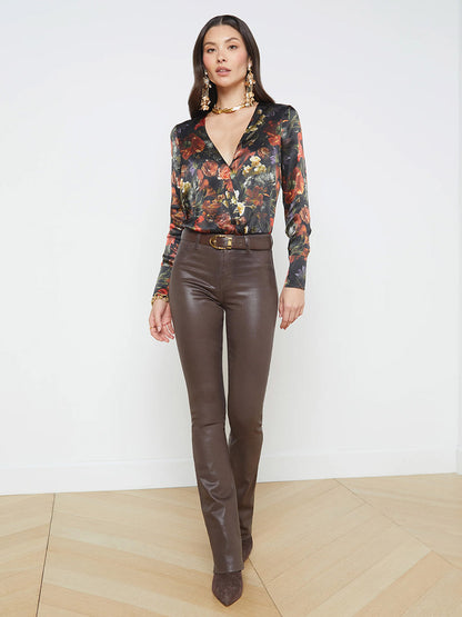 A person wearing a floral long-sleeve top, L'Agence Ruth High Rise Straight-Leg Jean in Espresso Coated, and large gold earrings stands on a wooden floor against a white wall. The straight-leg cut of the jeans subtly complements the overall chic ensemble.