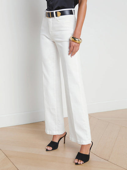 A woman in L'Agence Scottie wide-leg jeans in Blanc and black high-heeled sandals, accessorized with a black belt and gold bangle, standing in a minimalist room.