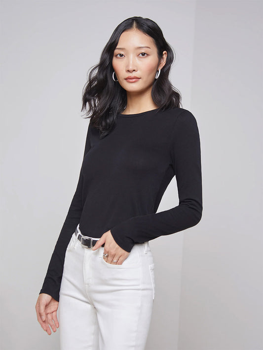 A person with long dark hair is wearing the L'Agence Tess Long Sleeve Crew in Black, white pants, and hoop earrings, standing against a plain background.