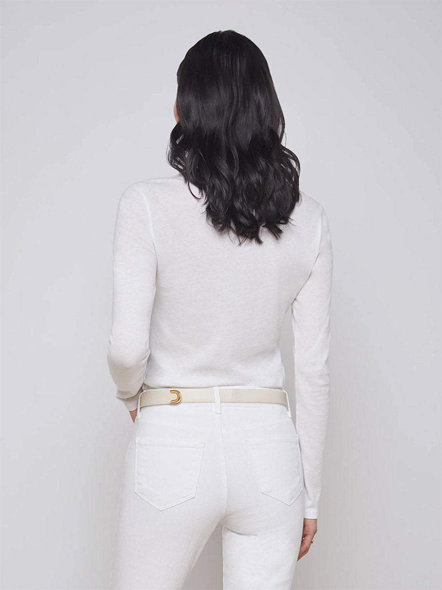 A person with shoulder-length dark hair is seen from the back, wearing the L'Agence Tess Long Sleeve Crew in White and white pants with a beige belt. The outfit, made in the USA, showcases simplicity and elegance.