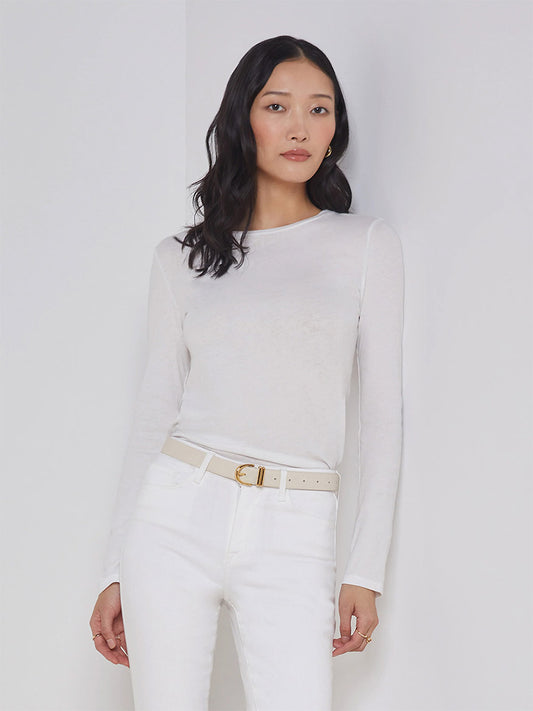 A person with long, dark hair is standing against a white background, wearing the L'Agence Tess Long Sleeve Crew in White and white pants paired with a beige belt.