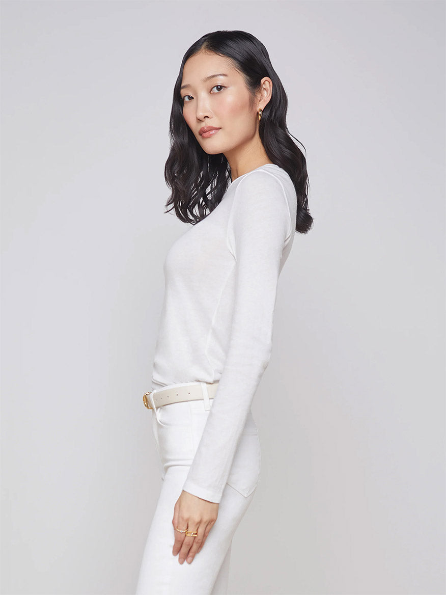 A woman with long dark hair is wearing the L'Agence Tess Long Sleeve Crew in White along with matching pants, standing against a plain light background. The center back length of the top is 25", and it proudly displays the "Made in USA" label.