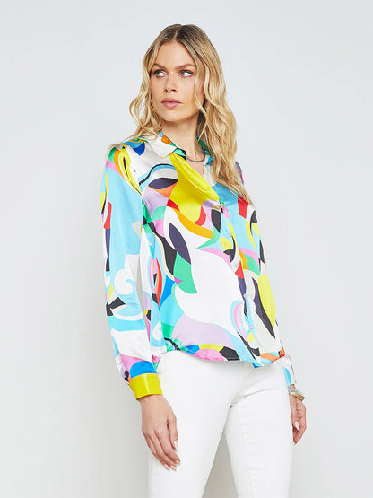 A woman with wavy blonde hair wears a L'Agence Tyler Blouse in Small Multi Kaleidoscope and white pants, standing against a plain background. This timeless design is perfect for adding flair to any versatile wardrobe.