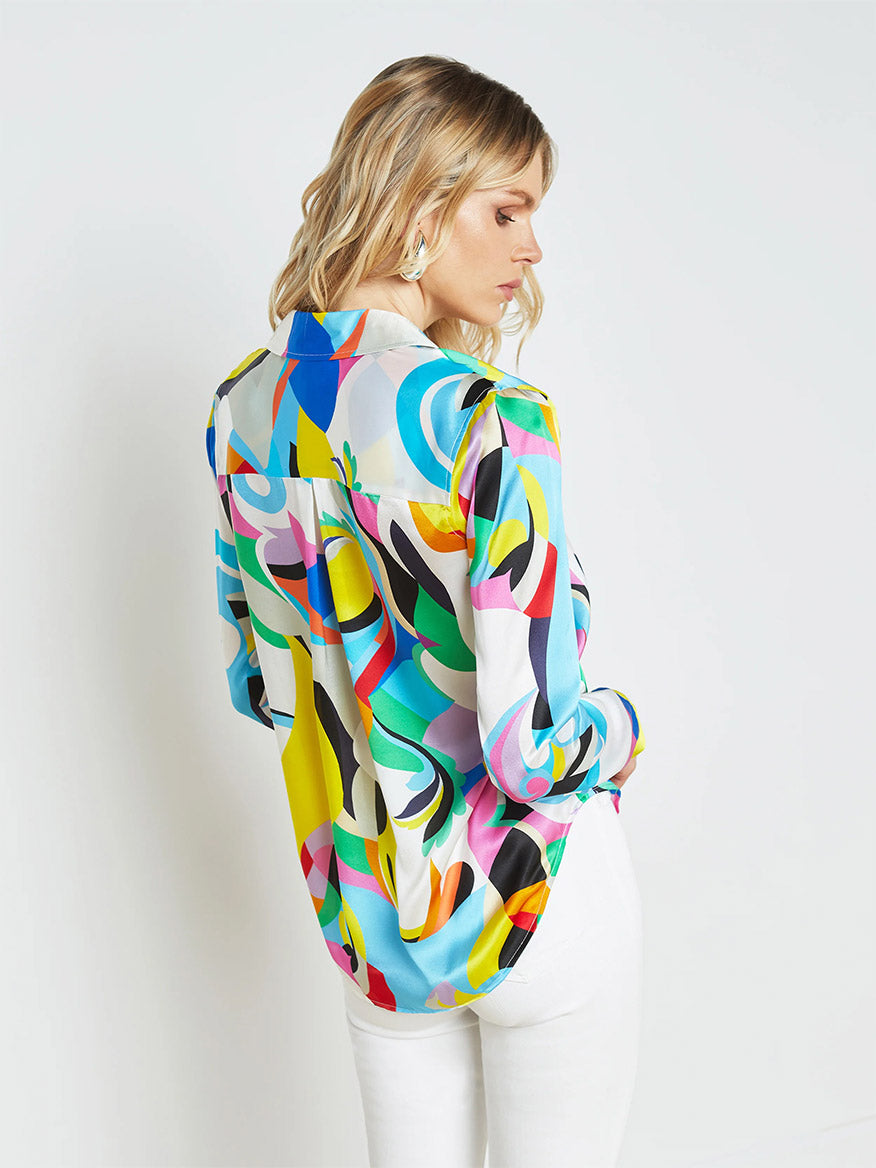 A person with blonde hair is turned away, wearing a colorful, patterned L'Agence Tyler Blouse in Small Multi Kaleidoscope and white pants, showcasing a versatile wardrobe against a plain white background.