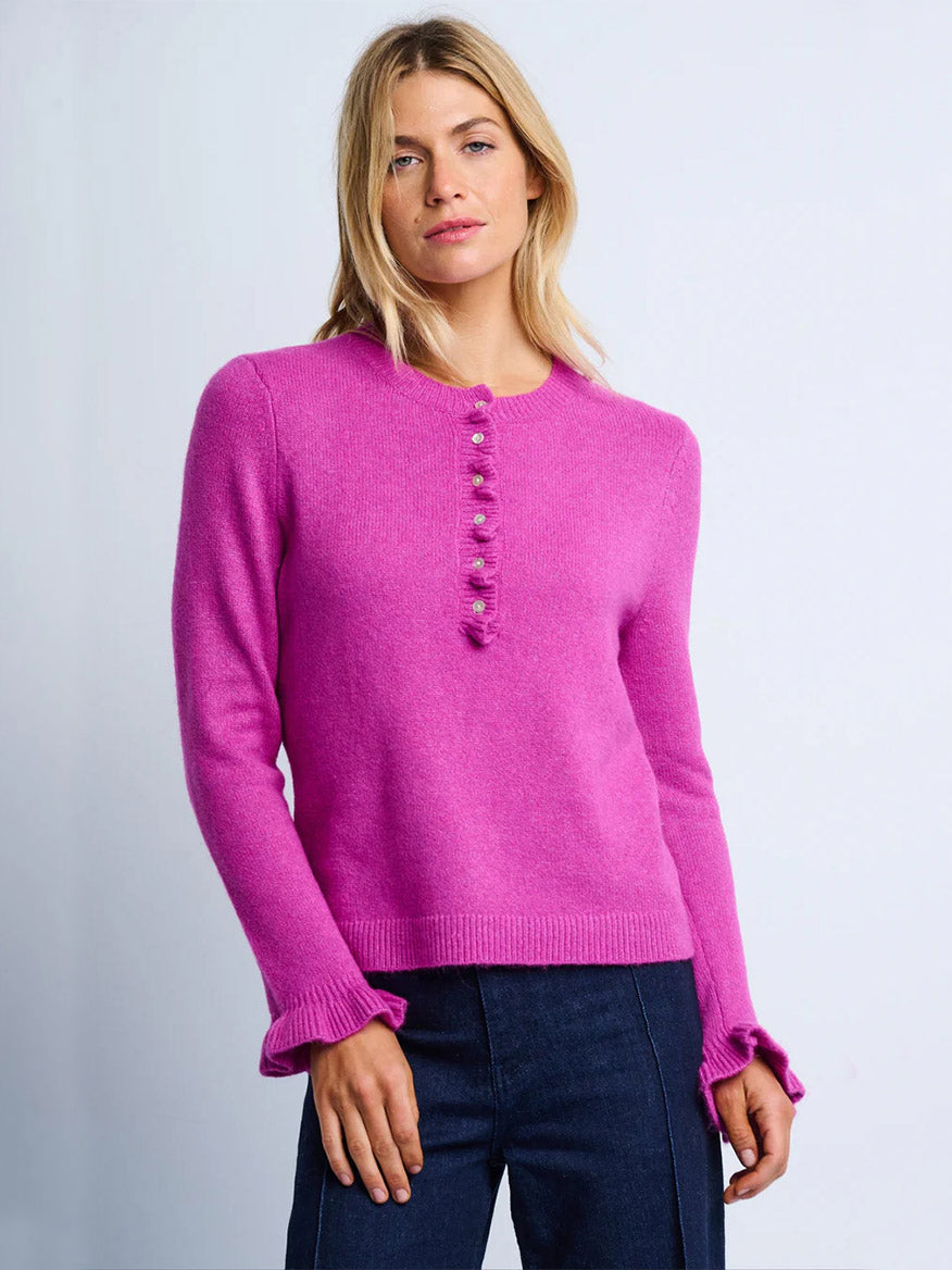 A woman stands against a plain background wearing the Lisa Todd Allure Henley in Hot Orchid, featuring a ruffled placket and decorative buttons, crafted from recycled fibers, paired with dark blue pants.