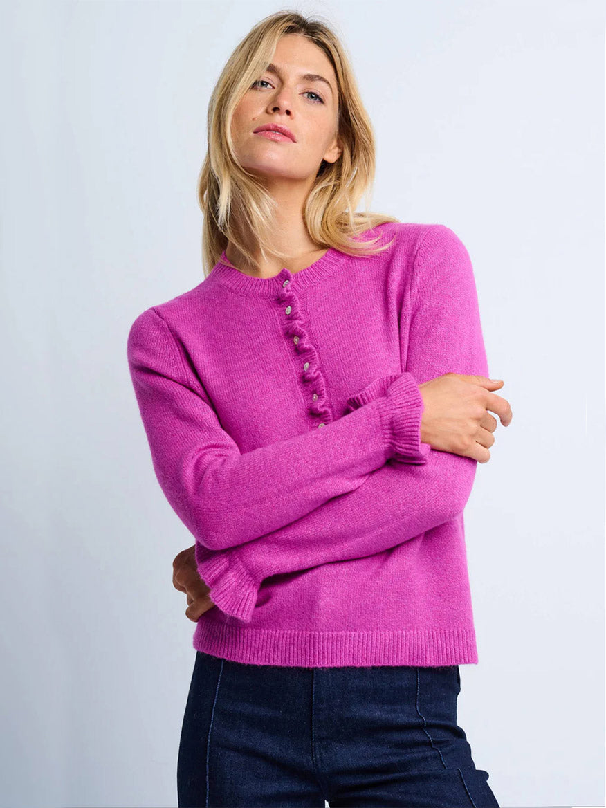 A woman wears the Lisa Todd Allure Henley in Hot Orchid, a bright pink long-sleeve sweater with decorative buttons and a ruffled placket, crafted from soft recycled fibers, standing against a plain light background.