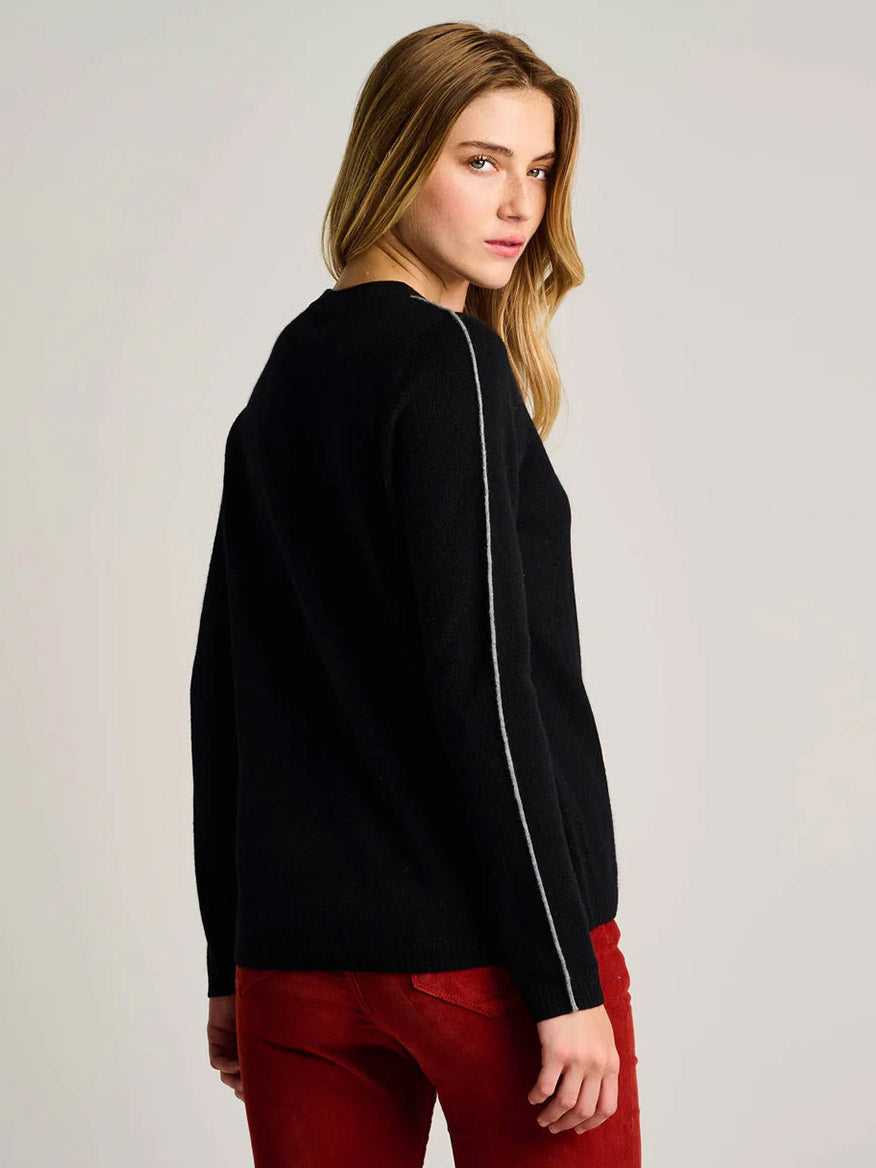 A woman with long blonde hair models the Lisa Todd Après All Day Sweater in Onyx—a black sweater with a white sleeve stripe—paired with red pants, standing and glancing over her shoulder.