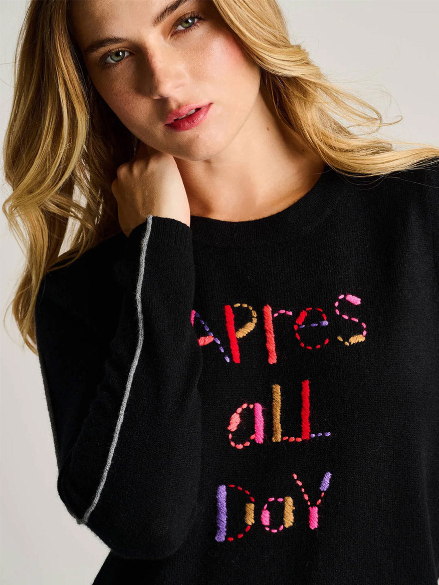 A blonde woman wears the Lisa Todd Après All Day Sweater in Onyx, a luxe black pullover featuring colorful "Après all day" text on the front.