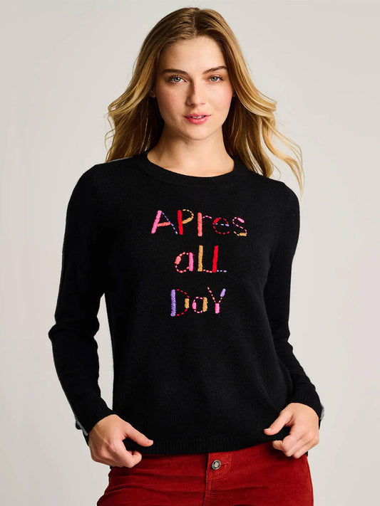 A woman wears the Lisa Todd Après All Day Sweater in Onyx, featuring colorful lettering, paired with red pants against a plain background.