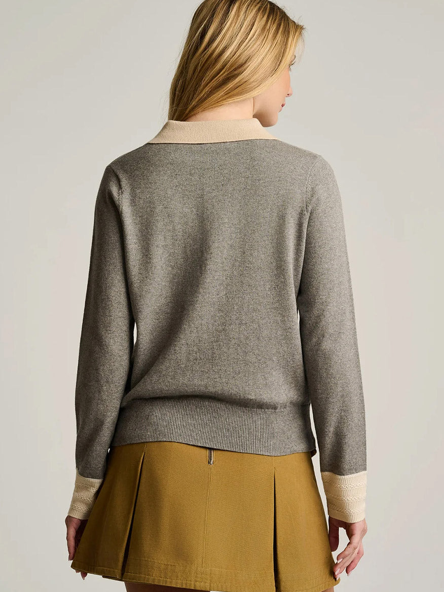 A woman with blonde hair is shown from the back, wearing the Lisa Todd Pop Posh Cotton Cashmere Polo in Grey Shadow, distinguished by its cream cuffs and collar, and paired elegantly with a mustard-colored pleated skirt.