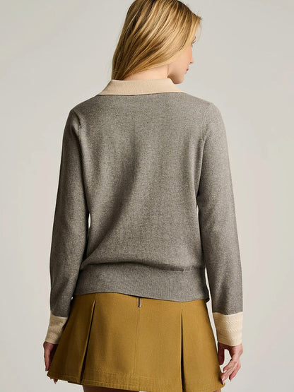 A woman with blonde hair is shown from the back, wearing the Lisa Todd Pop Posh Cotton Cashmere Polo in Grey Shadow, distinguished by its cream cuffs and collar, and paired elegantly with a mustard-colored pleated skirt.