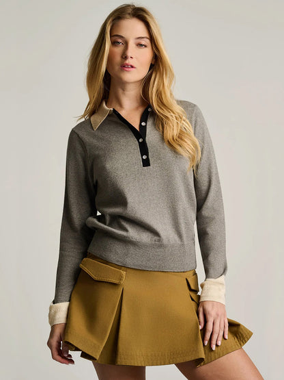 A woman stands against a plain background wearing the Lisa Todd Pop Posh Cotton Cashmere Polo in Grey Shadow, paired with a tan pleated skirt.