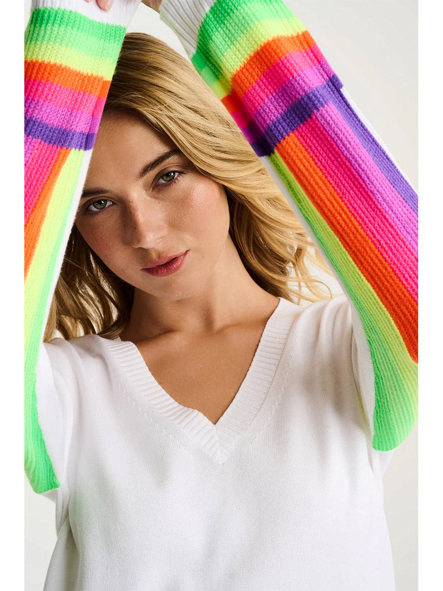 Wearing the Lisa Todd Brite Side Cotton Sweater in White, a woman raises her arms to partially frame her face and gazes at the camera, showcasing the sweater’s colorful rainbow-striped sleeves.