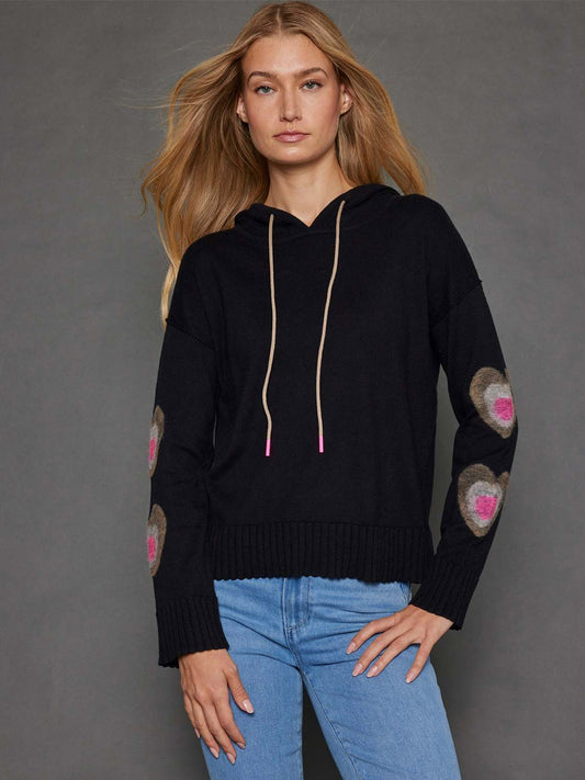 A person with long hair models the Lisa Todd Catch Me Hoodie in Black, featuring luxurious cashmere and bold hearts on the elbows, paired with blue jeans, posed against a gray background.