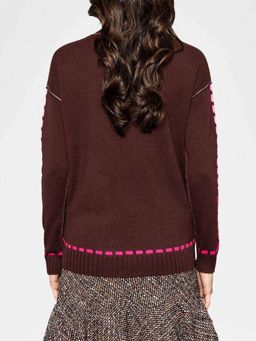 A person with long brown hair is wearing the Lisa Todd Dash Cam Sweater in Coco, featuring burgundy with contrast trim and pink accents, paired with a patterned skirt, seen from behind.