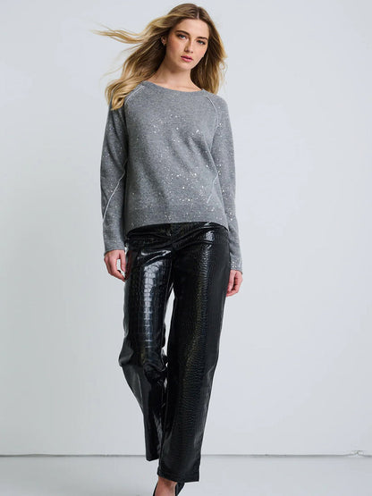 A woman stands against a plain background wearing the Lisa Todd Diamond Dust Sweater in Fog and black faux-leather pants with a textured pattern.