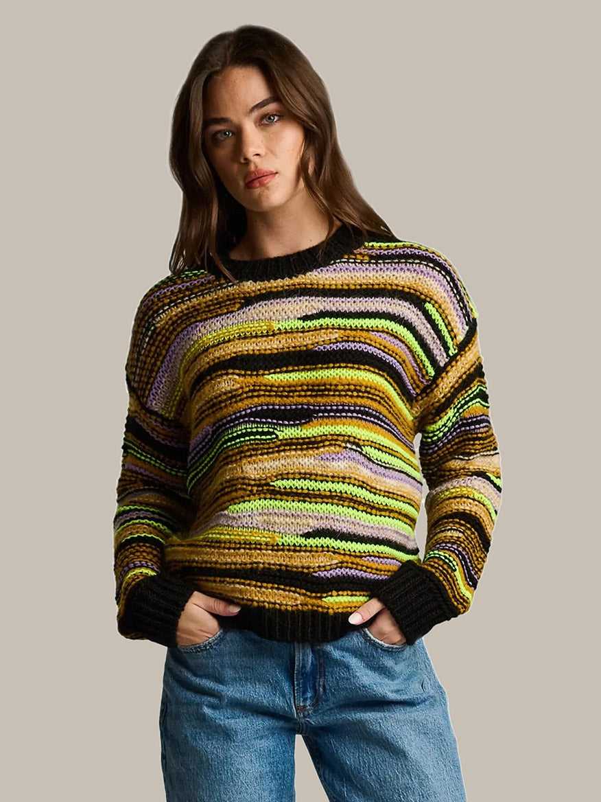 A woman wearing a black, yellow, and purple striped Lisa Todd Fillati Sweater in Black Multi, showcasing artisanal knit craftsmanship in a long-sleeve design.