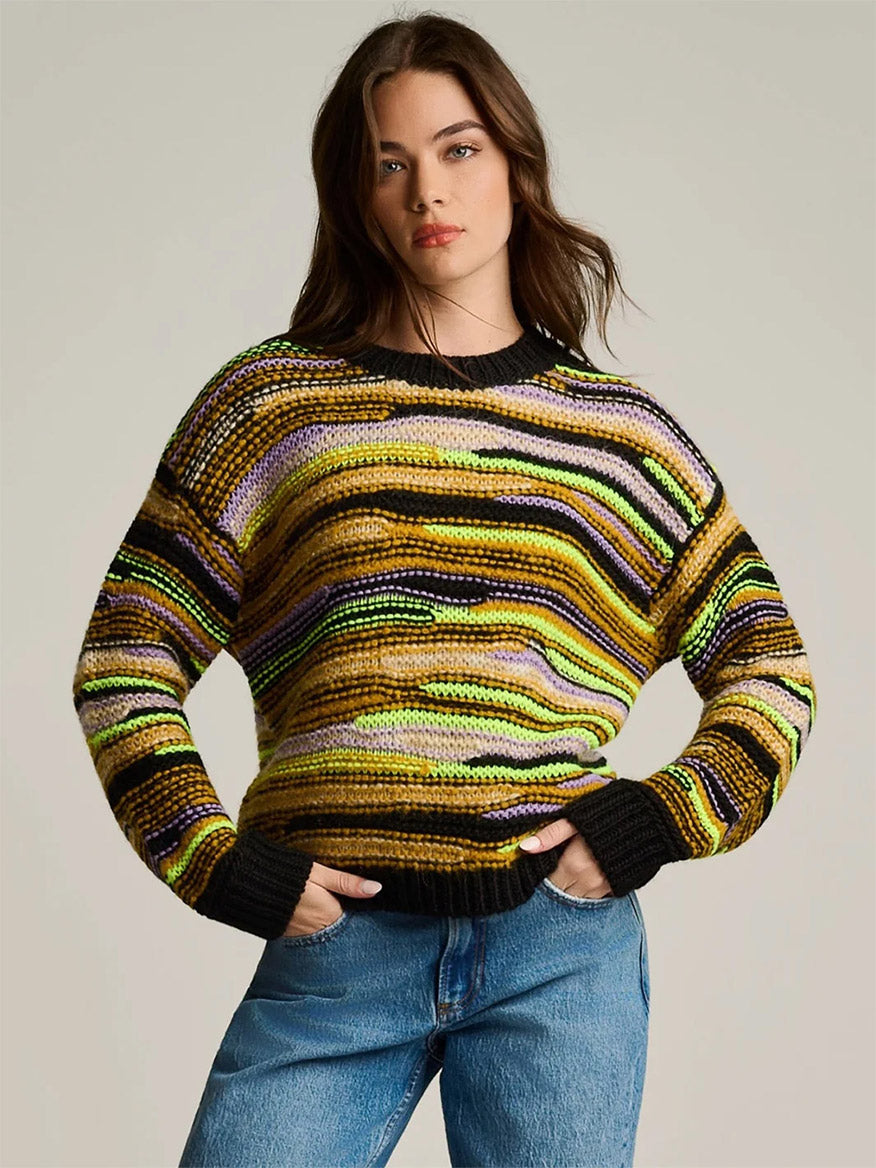 A person wears a Lisa Todd Fillati Sweater in Black Multi featuring horizontal stripes of black, lime green, tan, and purple, paired with blue jeans.