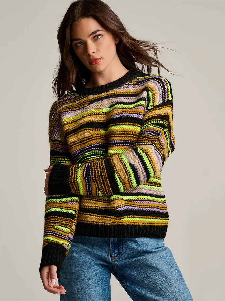 A woman wears a Lisa Todd Fillati Sweater in Black Multi, a ribbed knit sweater featuring horizontal stripes in black, yellow, purple, and cream, paired with blue jeans.