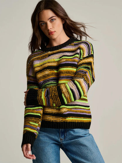 A woman wears a Lisa Todd Fillati Sweater in Black Multi, a ribbed knit sweater featuring horizontal stripes in black, yellow, purple, and cream, paired with blue jeans.