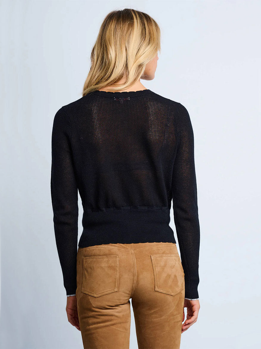 A blonde woman stands facing away, wearing the Lisa Todd Flirt Cardigan in Black and tan suede pants against a light background.