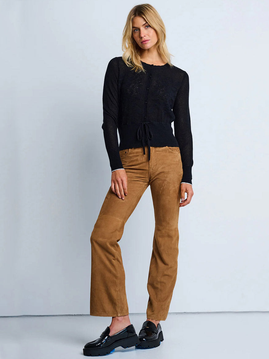 A woman stands against a plain background wearing the Lisa Todd Flirt Cardigan in Black, tan wide-leg pants, and black platform shoes.