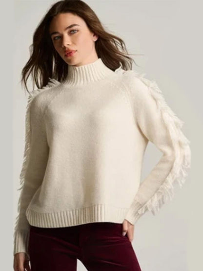 Woman wearing a Lisa Todd Fringe Benefits Sweater in Winter White—high-neck, relaxed raglan style with fringe—paired with maroon pants against a plain background.