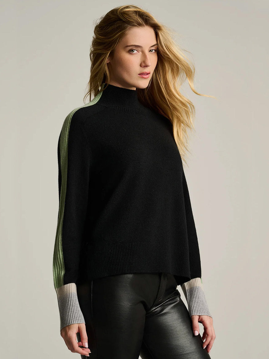 A woman wears the Lisa Todd High Spirits Mock in Onyx, a relaxed raglan silhouette black mock-neck sweater with wide, pale green stripes down the shoulders and color-blocked grey cuffs.