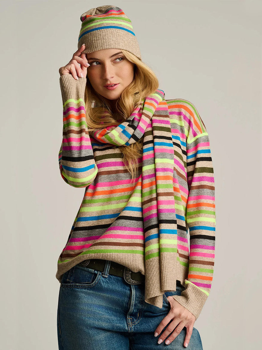 A woman stands against a plain background, wearing blue jeans and the Lisa Todd Hype The Stripe Hat in Oat Multi Stripe, paired with a matching striped sweater and scarf.