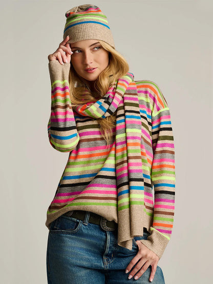 A woman stands against a plain background, wearing blue jeans and the Lisa Todd Hype The Stripe Hat in Oat Multi Stripe, paired with a matching striped sweater and scarf.