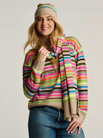 Woman wearing blue jeans, a colorful striped sweater, and the Lisa Todd Hype The Stripe Scarf in Oat Multi Stripe, smiling against a plain background.
