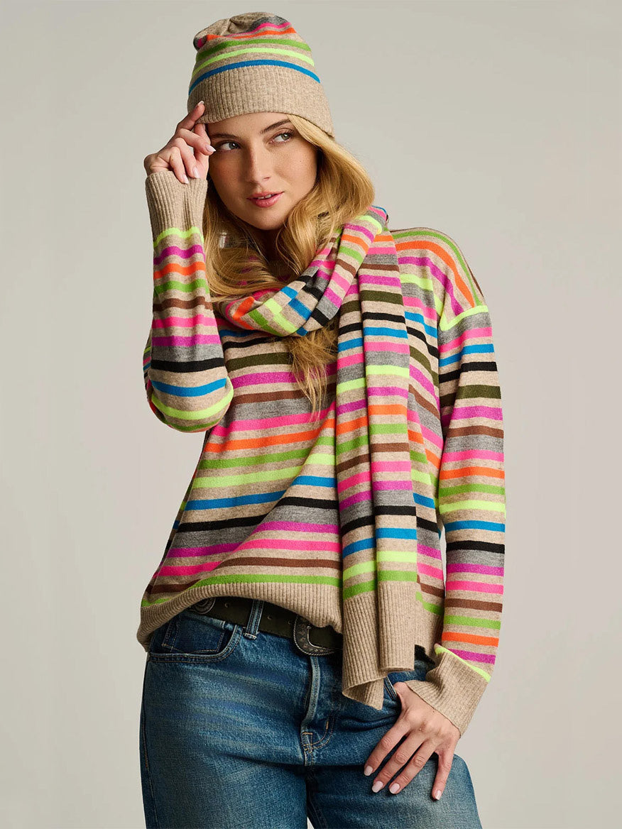 Woman wearing Lisa Todd Hype The Stripe Scarf in Oat Multi Stripe, a colorful sweater, knit hat, and blue jeans against a plain background.
