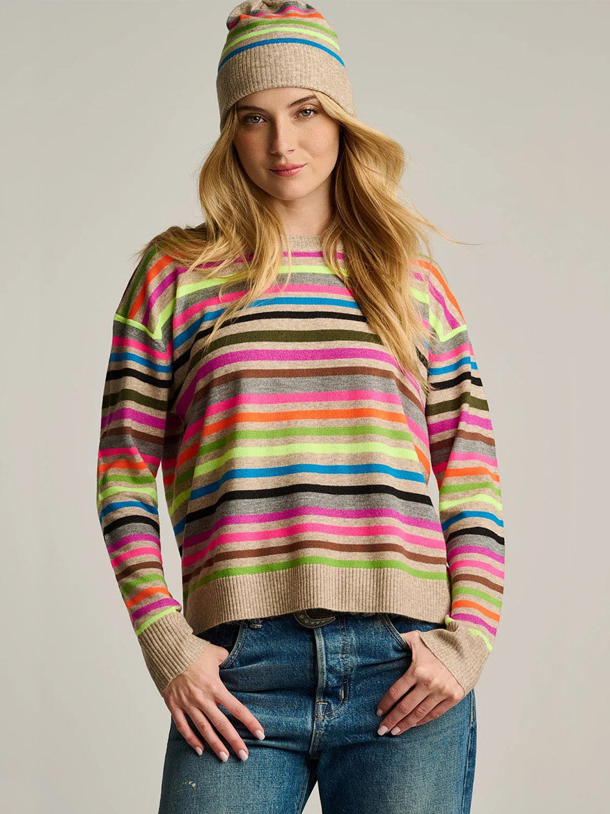Woman wearing the Lisa Todd Hype The Stripe Sweater in Oat Multi Stripe and matching beanie, paired with blue jeans against a plain background.
