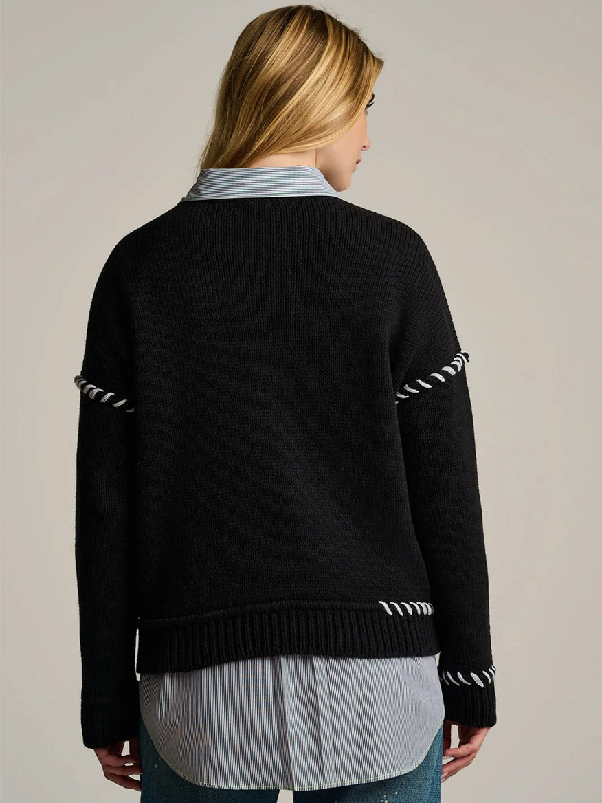 Person with long blonde hair, facing away, wears the Lisa Todd In Bloom Sweater in Black over a blue collared shirt and blue jeans against a plain background.