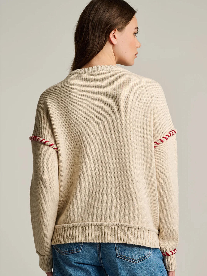 A person with long brown hair wears the Lisa Todd In Bloom Sweater in Salty, featuring red contrast stitching on the sleeves, and blue jeans, seen from behind.