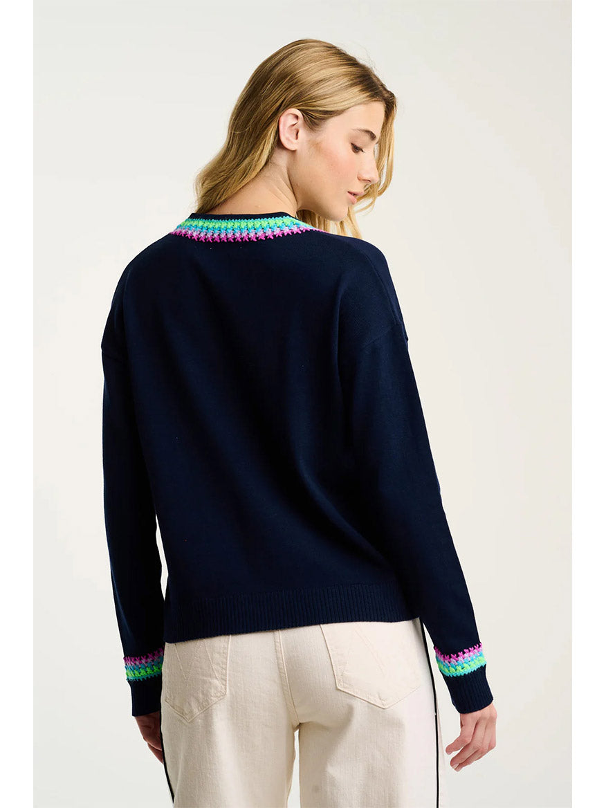 A woman with blonde hair, facing away from the camera, wears the Lisa Todd In The Mix Sweater in Navy featuring a relaxed V-neck and multicolored stitching at the collar and cuffs, paired with off-white pants.