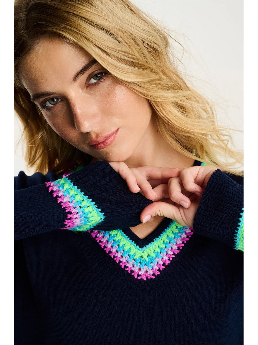 A woman with wavy blonde hair models the Lisa Todd In The Mix Sweater in Navy, featuring multicolored stitching on the neckline and cuffs, as she poses with her hands near her face.