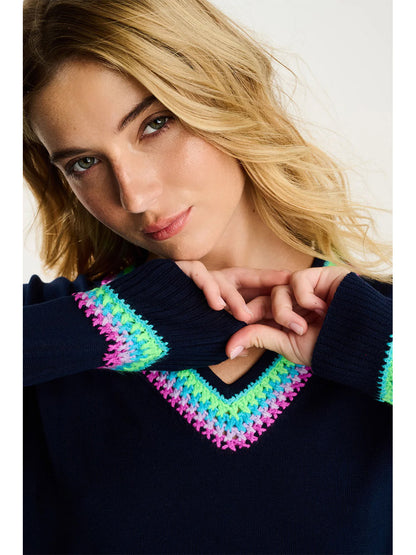 A woman with wavy blonde hair models the Lisa Todd In The Mix Sweater in Navy, featuring multicolored stitching on the neckline and cuffs, as she poses with her hands near her face.