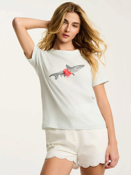 A woman poses against a plain background in the Lisa Todd Jaw-Dropping Tee in Salty Air, featuring a shark graphic with a pink bow, paired with white scalloped-hem shorts for a playful summer look.