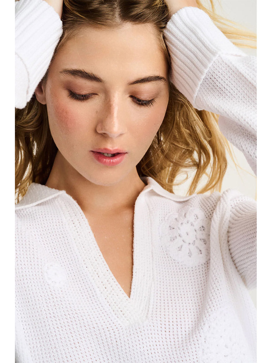 A blonde woman looks downward, wearing the Lisa Todd Knit Bliss Cotton Sweater in White, a textured 100% cotton V-neck with subtle floral patterns.