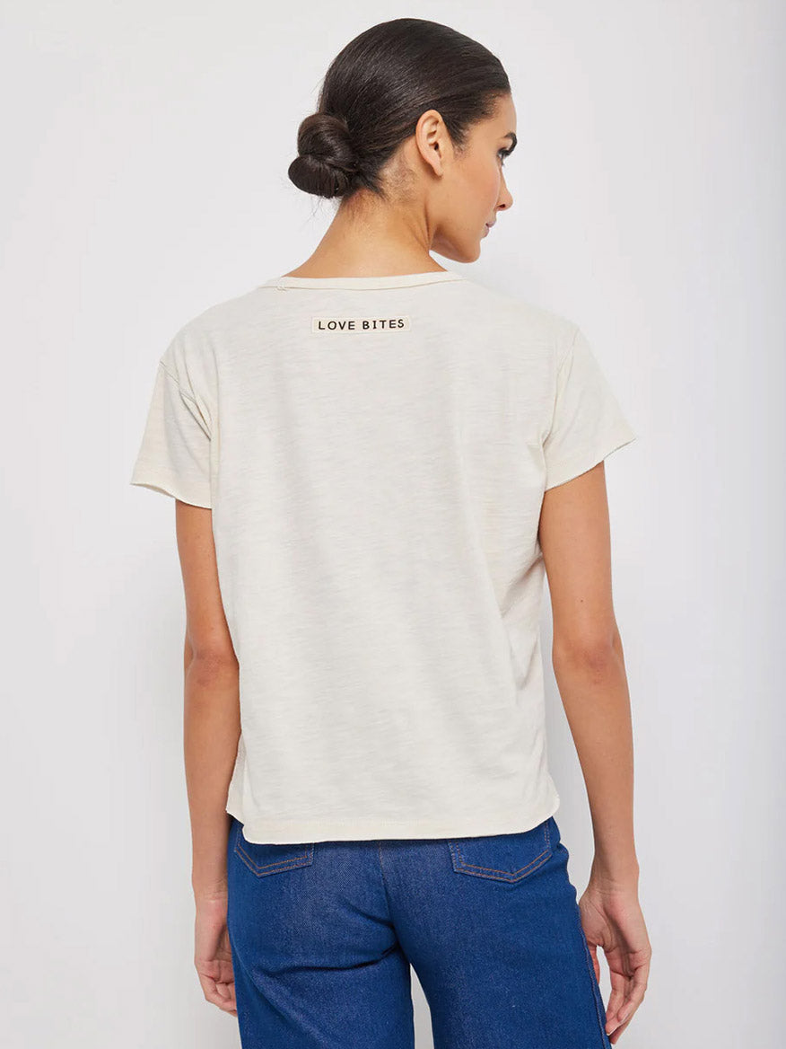 Woman seen from the back wearing a beige Lisa Todd Love Bites Tee in Bluff with "Love Bites" printed on the upper back area, paired with blue jeans featuring raw hems.