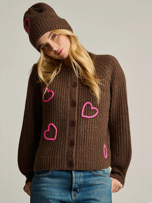 A woman wears the Lisa Todd Love Game Cardigan in Cocoa, an oversized, cozy brown ribbed knit cardigan and matching beanie, both featuring hand-embroidered neon pink heart outlines.