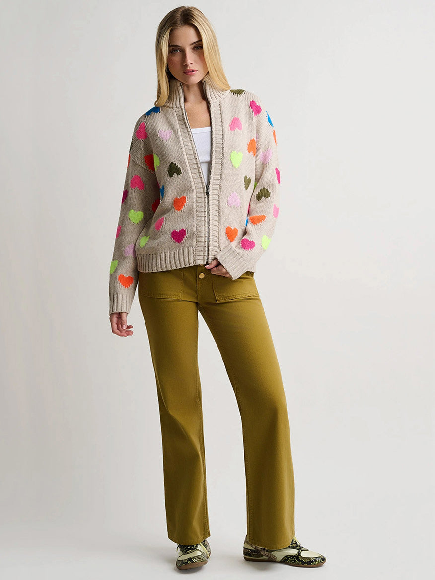 Lisa Todd Love Zip Cardigan in Chickpea featuring multicolored heart patterns, worn with olive green trousers and patterned loafers.