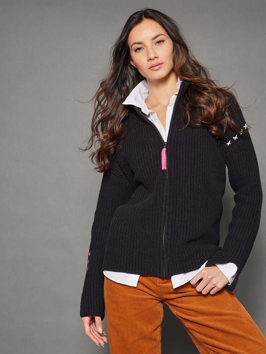 A person with long hair wearing the Lisa Todd Make Your Mark Zip Sweater in black over a white collared shirt and brown pants, standing against a gray background.