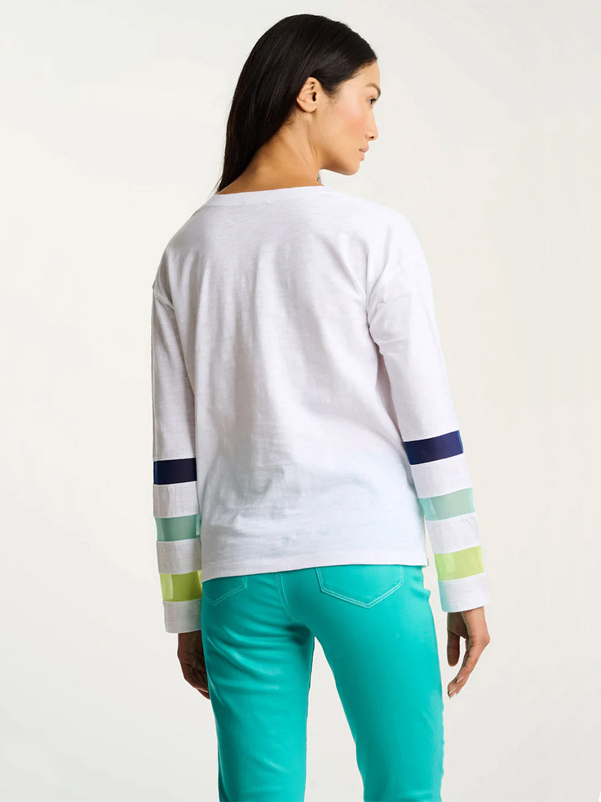 Woman stands with her back to the camera, wearing the Lisa Todd Mesh Fest Cotton Tee in White with colored stripes on the sleeves and teal pants.
